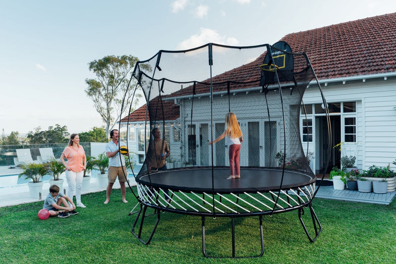 Family in a sunny backyard playing around a trampoline with a safety enclosure and basketball hoop