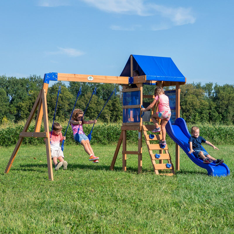 Children playing on a wooden swing set with a blue canopy, two belt swings, and a blue wave slide on a grassy lawn