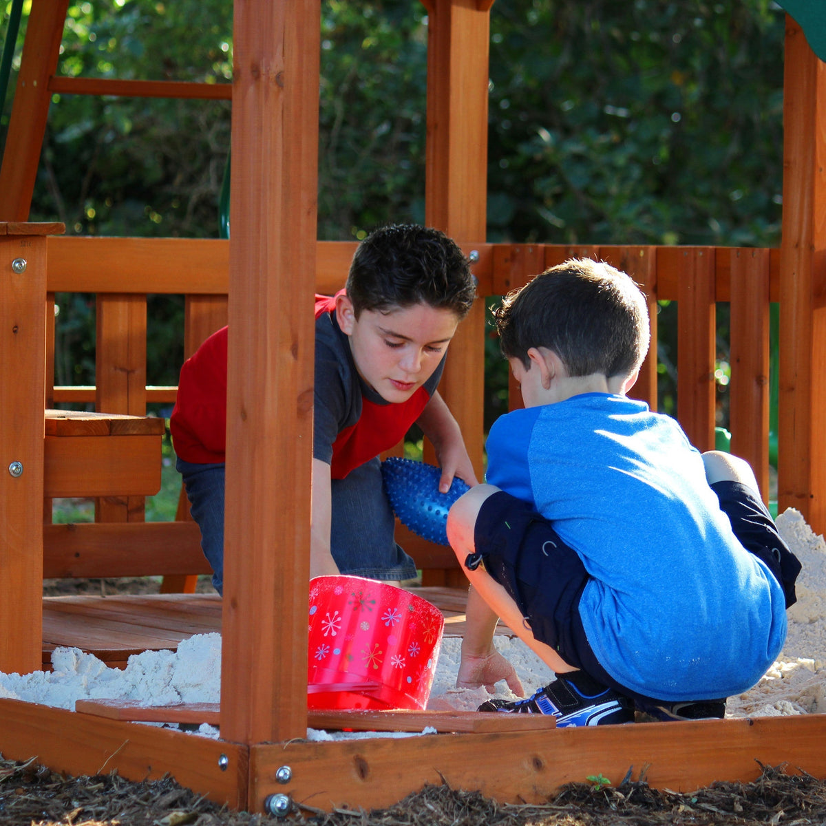 Two boys kneeling in a wooden sandbox beneath a cedar playset deck, scooping sand with a red bucket