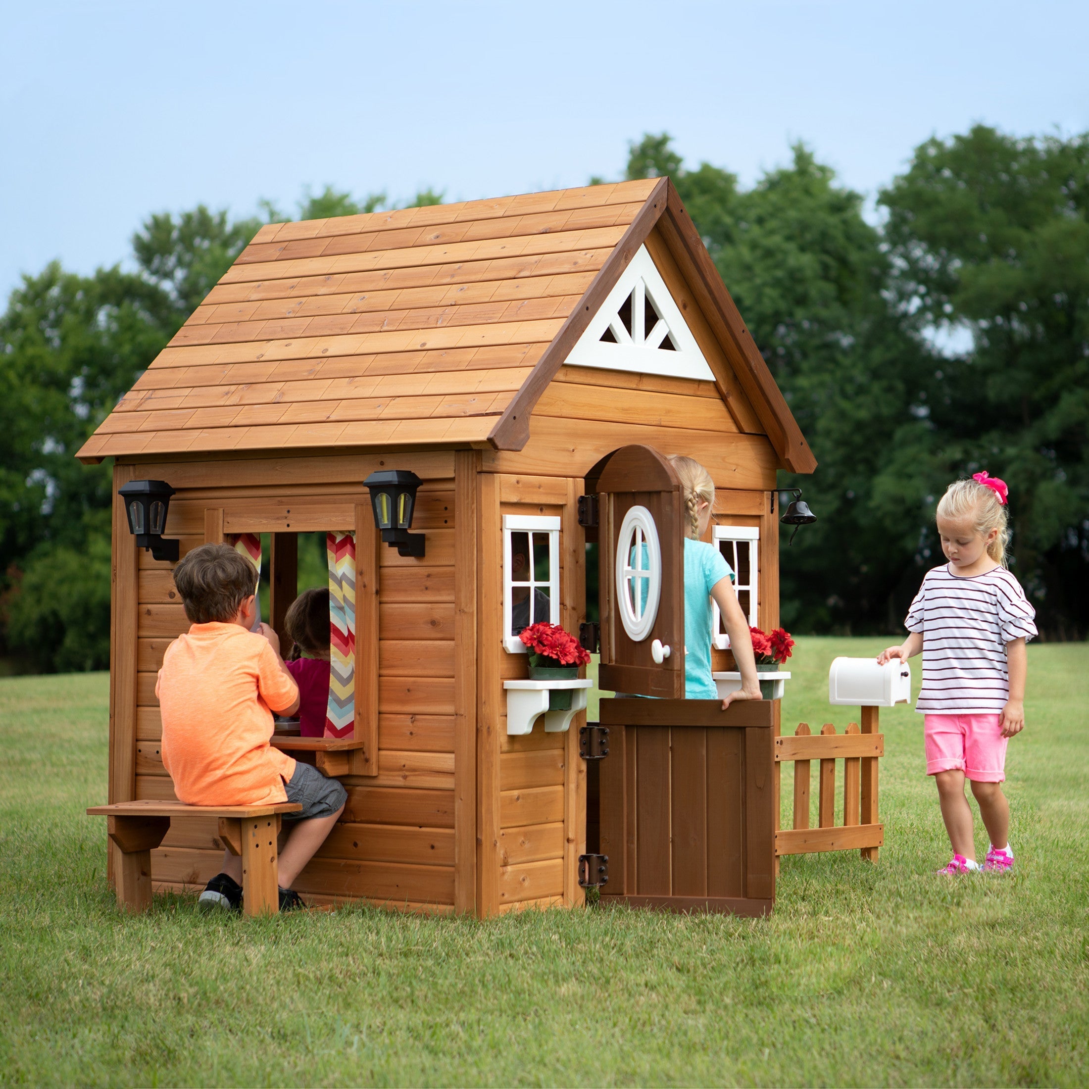 Children playing around a wooden outdoor playhouse with a pitched roof, windows, flower boxes, and a small front fence in a grassy backyard
