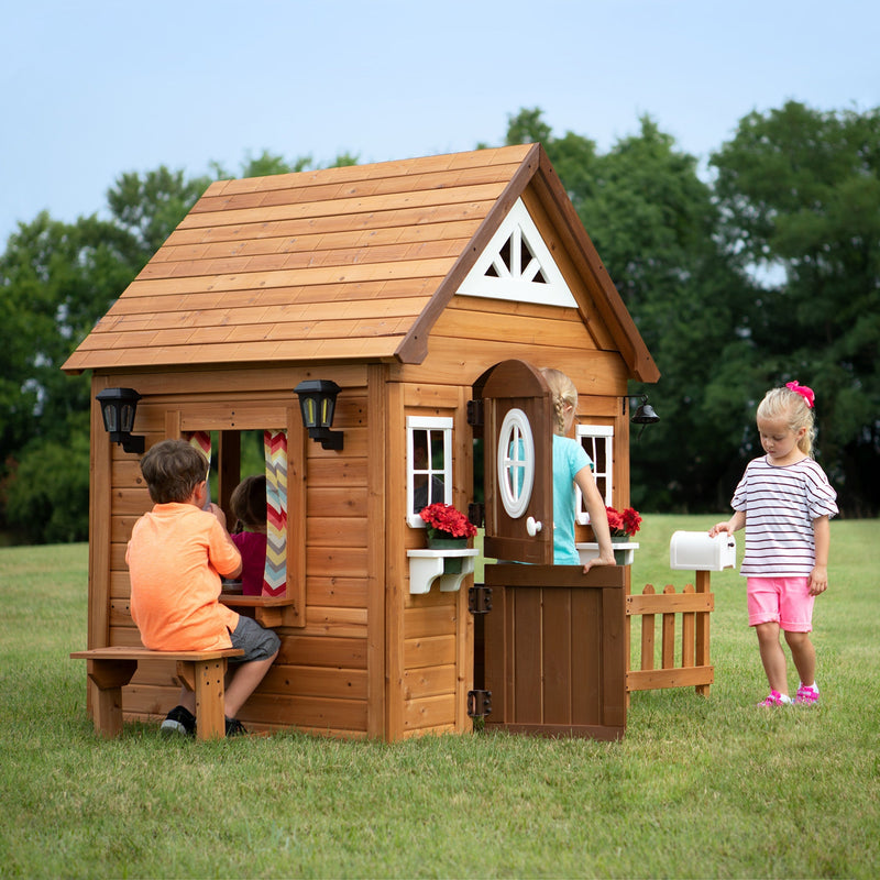 Children playing around a wooden outdoor playhouse with a pitched roof, windows, flower boxes, and a small front fence in a grassy backyard