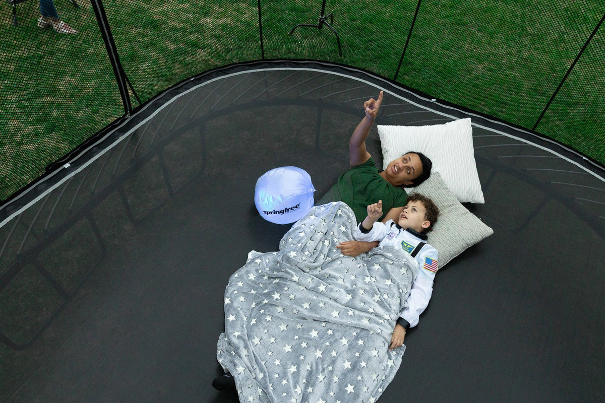 Parent and child lying under a blanket on a Springfree trampoline mat, pointing up at the sky
