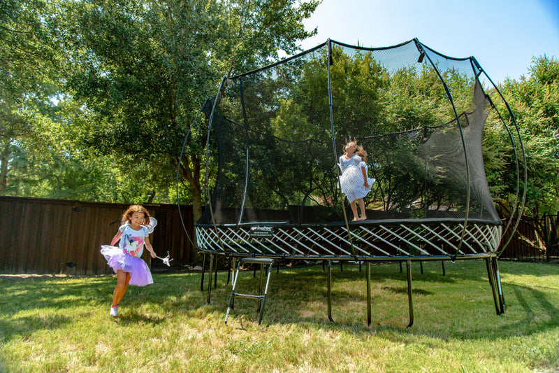 Child jumping on a round backyard trampoline with a safety enclosure while another child runs across the lawn