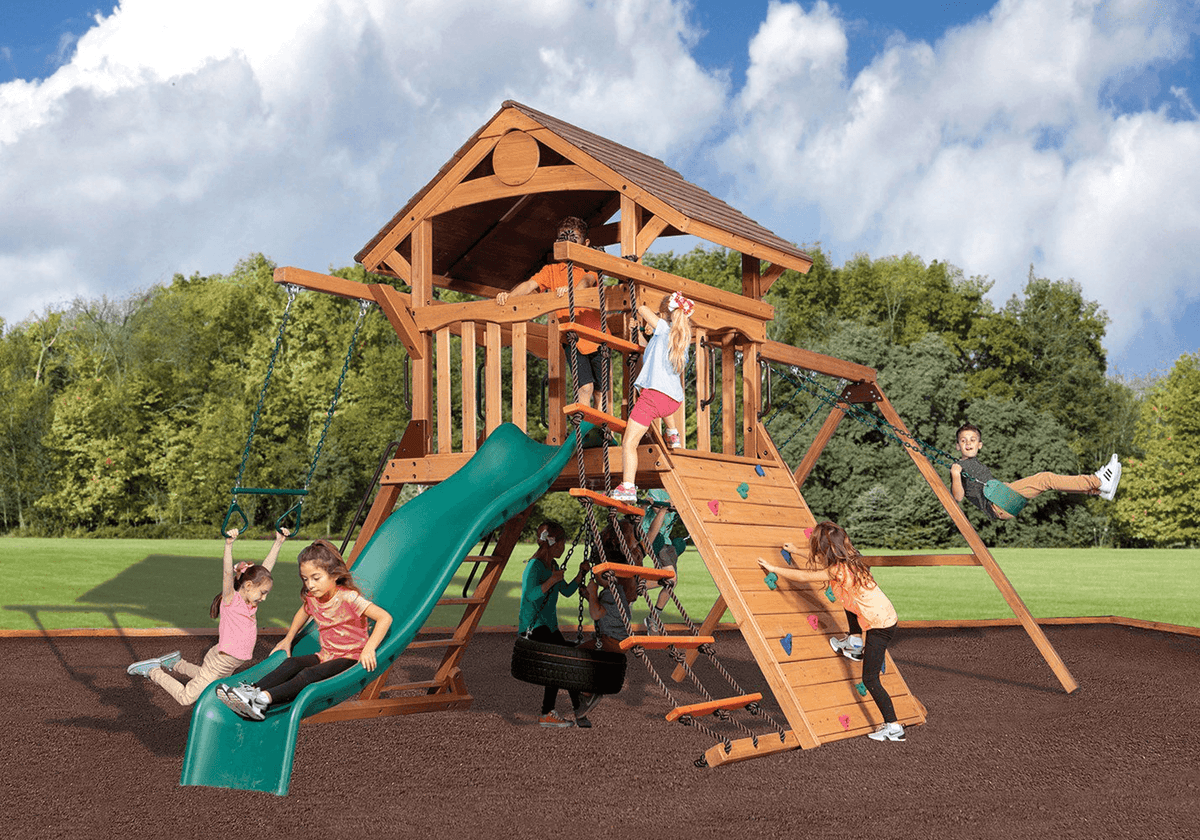 Kids playing on a cedar playset with a green wave slide, climbing wall, tire swing and rope ladder over rubber mulch