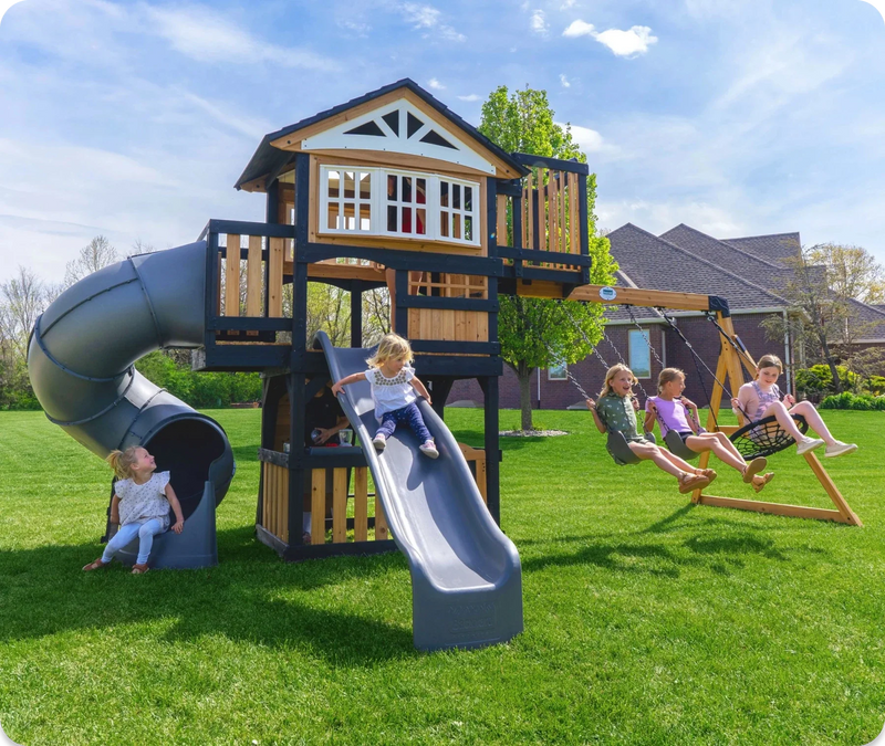 Kids sliding down a tube slide and wave slide and swinging on a cedar playset set on a flat green lawn