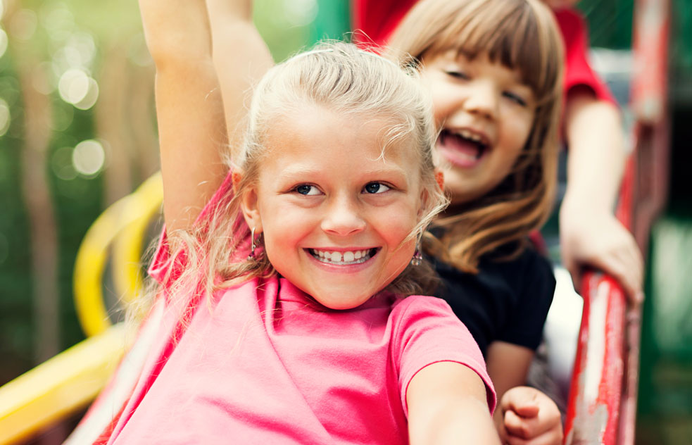 Two smiling girls playing together on an outdoor playground slide on a sunny day