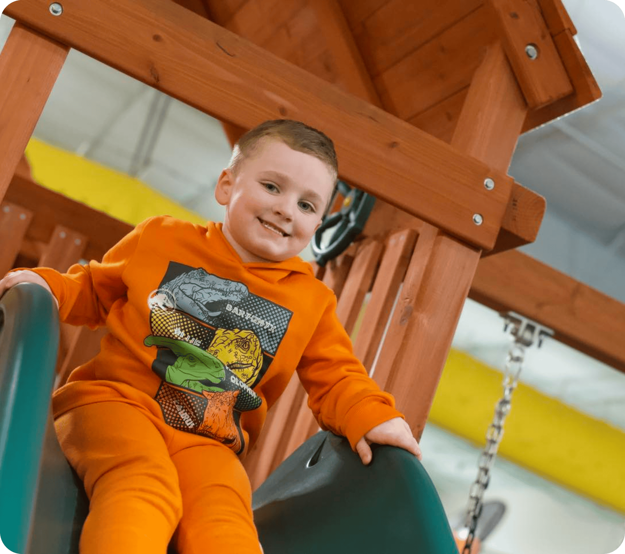 Young boy in an orange sweatsuit smiling at the top of a slide on an indoor wooden playset