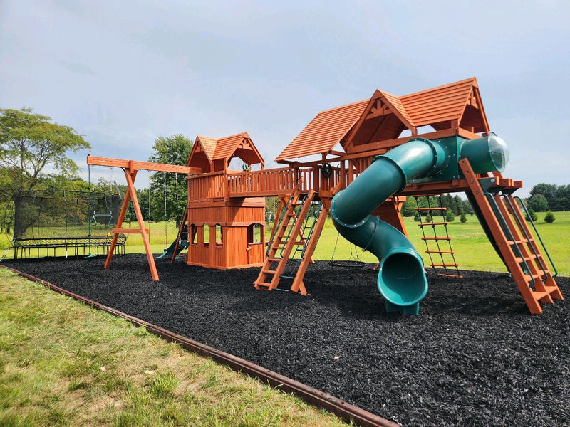 Large cedar playset with a green tube slide installed over a bed of black rubber mulch with an edged border