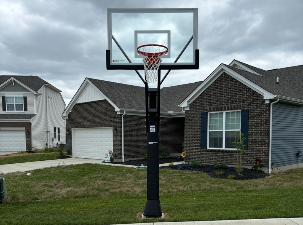 In-ground basketball hoop with a glass backboard on a black pole beside a driveway in front of a brick home