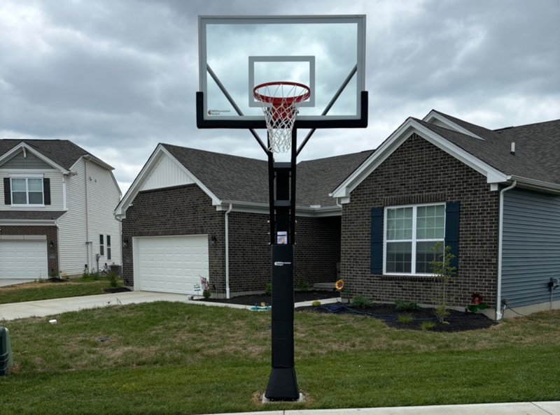 In-ground basketball hoop with a glass backboard on a black pole beside a driveway in front of a brick home