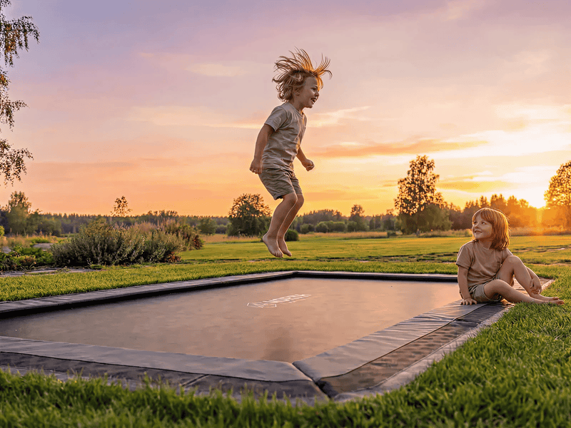Child jumping on an in-ground trampoline set flush with the lawn at sunset while another child sits on the edge