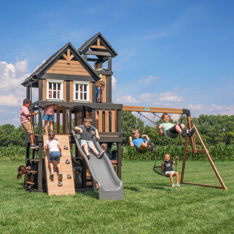 Kids climbing, sliding, and swinging on a wooden swing set with a two-story clubhouse, rock wall, and gray slide
