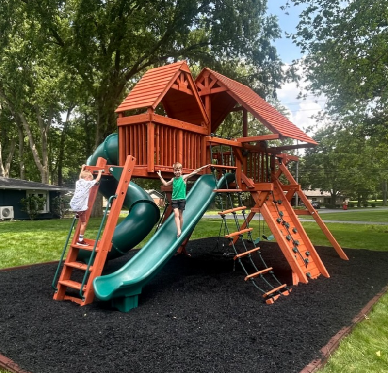 Two boys climbing a stained cedar playset with red-brown roofs, tube and wave slides, and a rope ladder over black rubber mulch