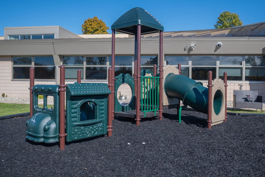 Green playground with a tunnel slide on rubber mulch in front of a school